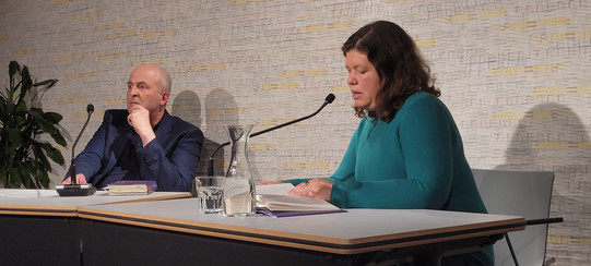 Das Bild zeigt Berthold Vogel links an einem gemeinsamen Tisch mit Julia Friedrichs (rechts) sitzen, während Friedrichs in ein Mikrofon spricht