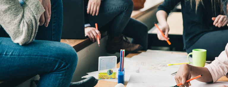 Brainstorming Vier Menschen sitzen an einem Tisch, die Gesichter sind nicht im Bild. Sie halten Stifte in der Hand und sehen nachdenkend aus. Eine Hand schreibt etwas auf das Plakat auf dem Tisch, welches sehr unstrukturiert gestaltet ist. Auf dem Tisch sind noch mehr Stifte und eine Tasse. Auf der Sitzbank erkennt man einen geöffneten Laptop und einen Rucksack im Hintergrund.